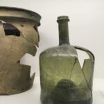 Zwei rekonstruierte Glasgefäße aus der Dauerausstellung im Museum: links ein braun-grünlicher Keramiktopf mit großen Fehlstellen, rechts eine grünliche Glasflasche mit sichtbaren Bruchkanten und ergänzten Fragmenten.