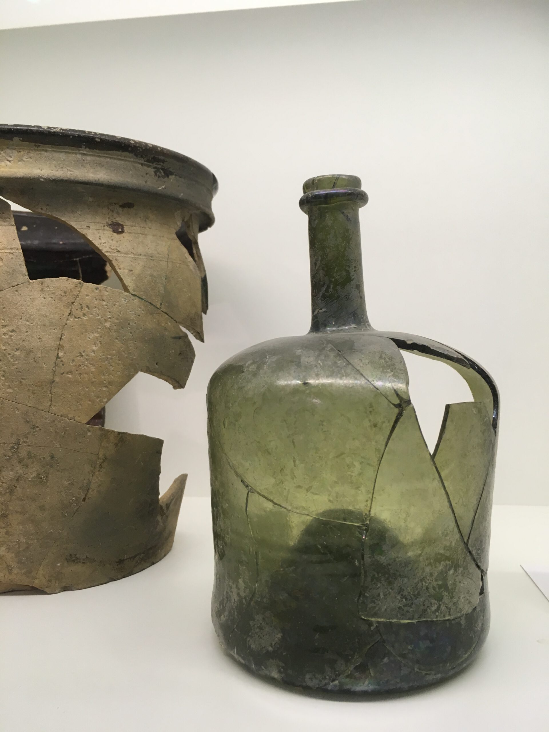 Zwei rekonstruierte Glasgefäße aus der Dauerausstellung im Museum: links ein braun-grünlicher Keramiktopf mit großen Fehlstellen, rechts eine grünliche Glasflasche mit sichtbaren Bruchkanten und ergänzten Fragmenten.