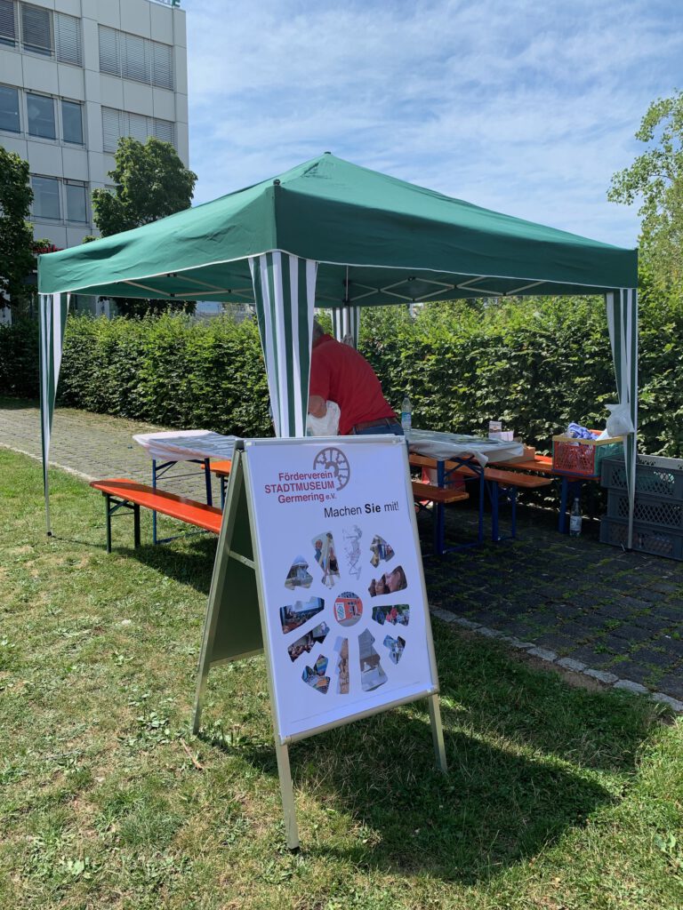 Infostand des Fördervereins Stadtmuseum Germering beim Stadtfest. Unter einem grünen Pavillon modellieren Kinder mit Ton kleine Gefäße. Im Vordergrund steht ein Aufsteller mit dem Logo des Fördervereins und der Aufschrift „Machen Sie mit!“.