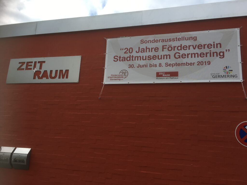 Au&szlig;enansicht des Museums ZEIT+RAUM in Germering mit roter Backsteinwand. Links das Museumsschild &bdquo;ZEIT + RAUM&ldquo;, rechts ein Banner zur Sonderausstellung &bdquo;20 Jahre F&ouml;rderverein Stadtmuseum Germering&ldquo; vom 30. Juni bis 8. September 2019.