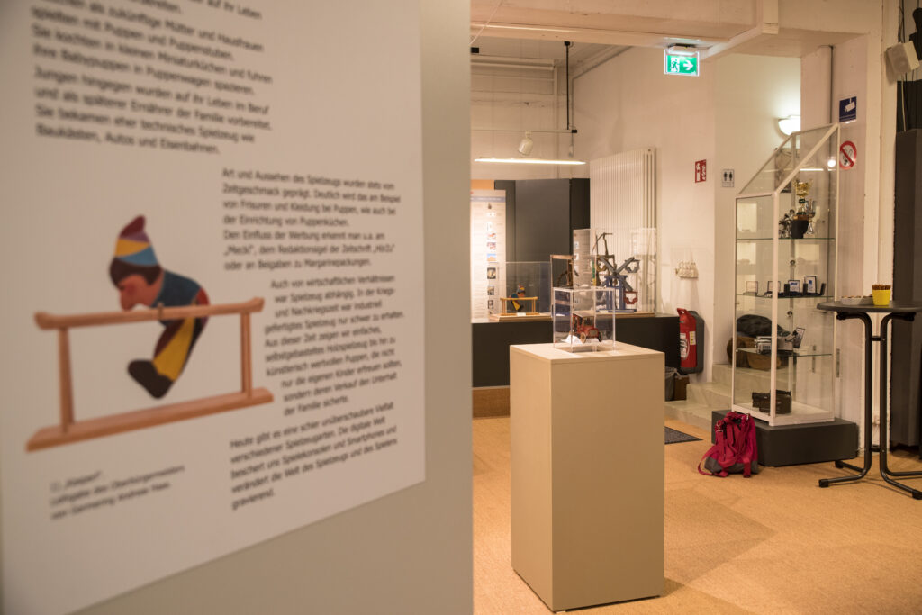 Blick in den Ausstellungsraum mit Vitrinen der Spielzeugausstellung. Im Vordergrund eine Texttafel mit einem Abbild des hölzernen „Kasperl“-Turnspielzeugs, im Hintergrund weitere Exponate, darunter ein Modellauto in einer Glashaube.