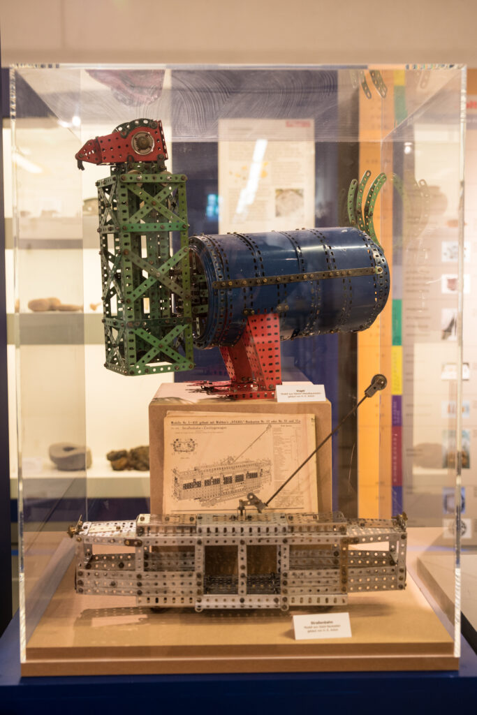Zwei Modelle aus Metallbaukästen in einer Vitrine im Museum: Unten ein silberner Straßenbahnwagen mit Stromabnehmer, darüber ein farbenfroher Vogel aus grünen, blauen und roten Metallteilen. Dazwischen eine historische Bauanleitung.