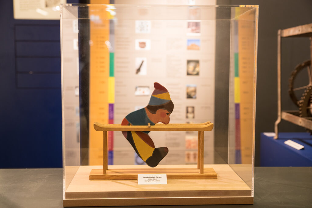 Holzspielzeug in Form eines bunten Kasperls, der als Turner an einer Reckstange befestigt ist. Ausgestellt in der Spielzeugausstellung.