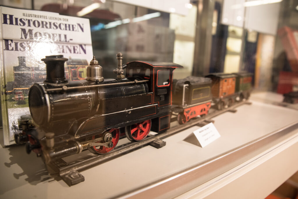 Vitrine mit einer historischen Spielzeugeisenbahn aus Blech, bestehend aus Lokomotive und mehreren Waggons, gezeigt in der Spielzeugausstellung im Museum. Im Hintergrund ein Buch mit dem Titel „Illustriertes Lexikon der historischen Modelleisenbahnen“, das die Präsentation ergänzt. Die Lokomotive ist auf einer Schiene montiert und mit auffälligen roten Speichenrädern ausgestattet.