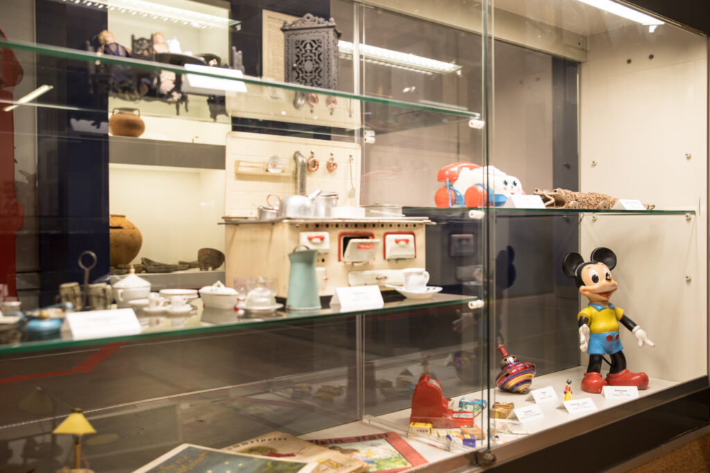 Vitrine in der Spielzeugausstellung im Museum mit historischem Kindergeschirr, einer Puppenküche, Blechspielzeug, einem Kreisel sowie einer großen Mickey Maus Figur.