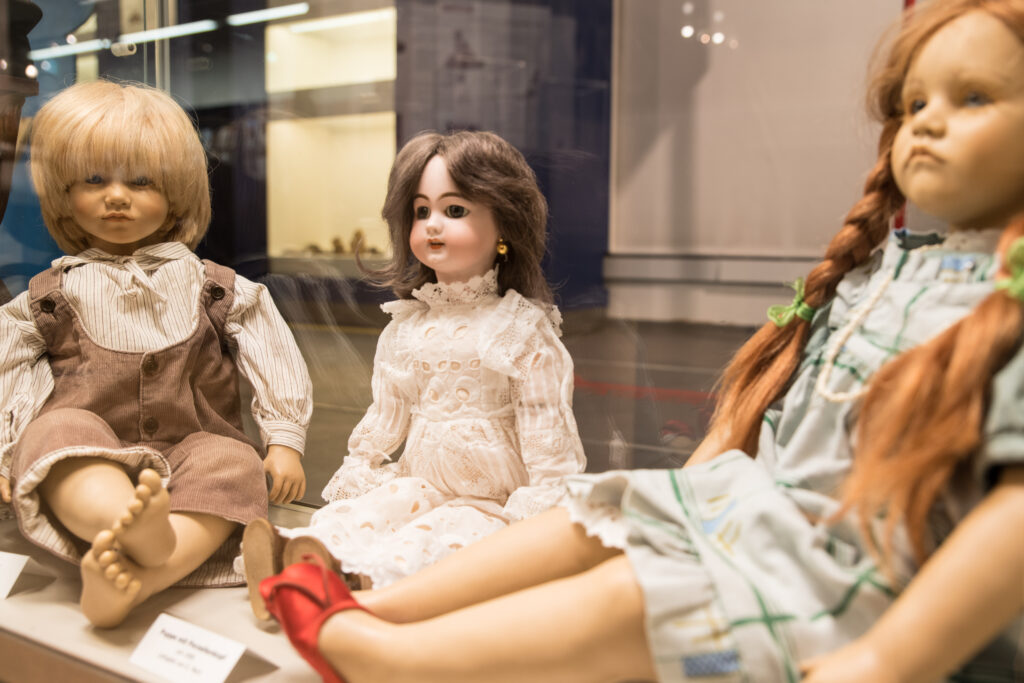 Vitrine mit drei verschiedenen historischen Puppen in der Spielzeugausstellung im Museum ZEIT+RAUM Germering. Zu sehen sind zwei moderne Künstlerpuppen sowie eine Porzellanpuppe in weißem Spitzenkleid aus dem frühen 20. Jahrhundert.