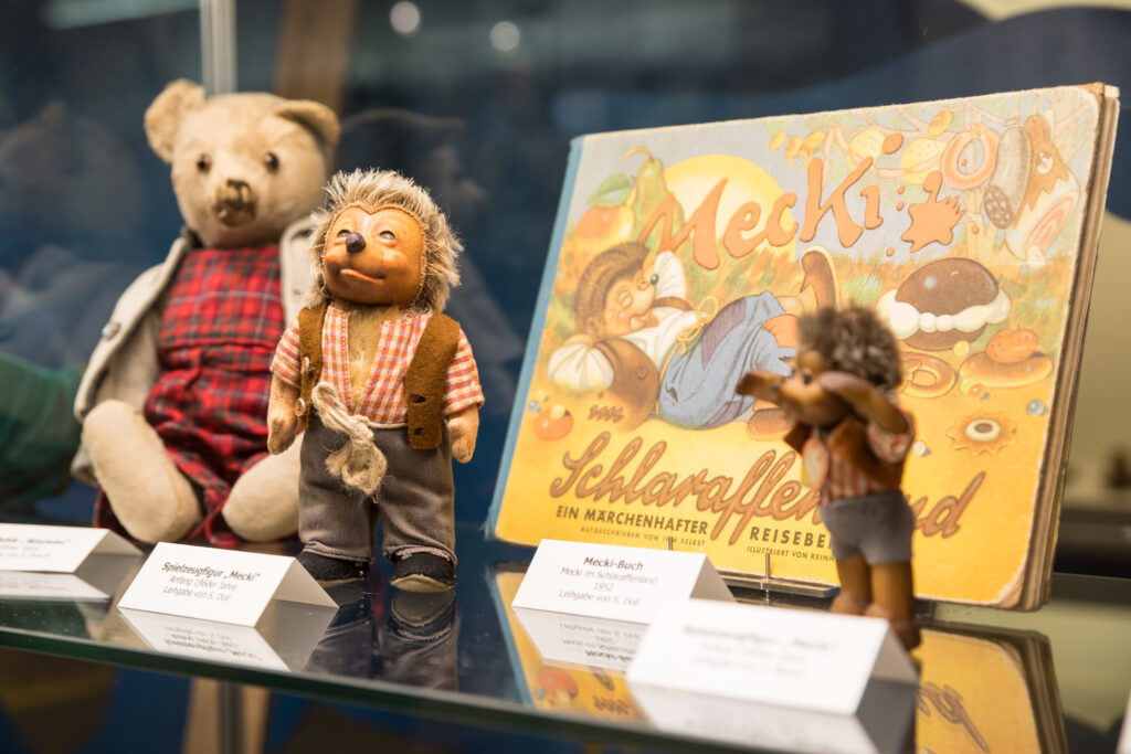 Vitrine mit Spielzeugfiguren und einem Kinderbuch aus der Spielzeugausstellung. Zu sehen ist unter anderem eine Mecki-Figur aus den 1950er-Jahren, das Buch „Mecki im Schlaraffenland“ sowie weitere passende Figuren.