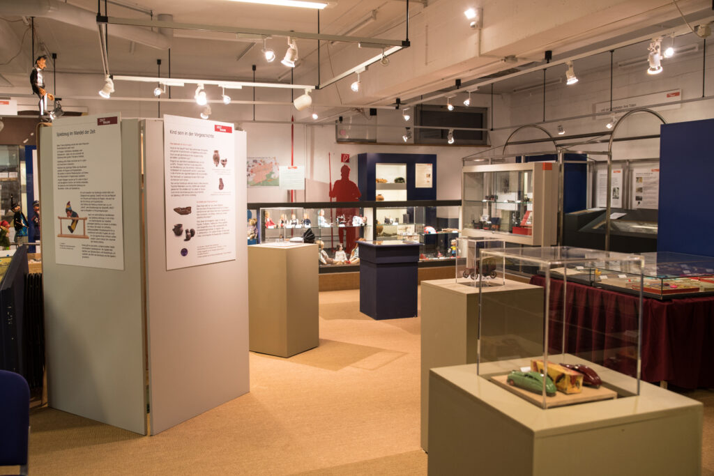 Ausstellungsraum der Spielzeugausstellung im Museum ZEIT+RAUM Germering mit zahlreichen Vitrinen, Schautafeln und historischen Spielzeugen. Im Vordergrund sind Blechspielautos in einer Glasvitrine zu sehen, im Hintergrund weitere Exponate und Figuren.