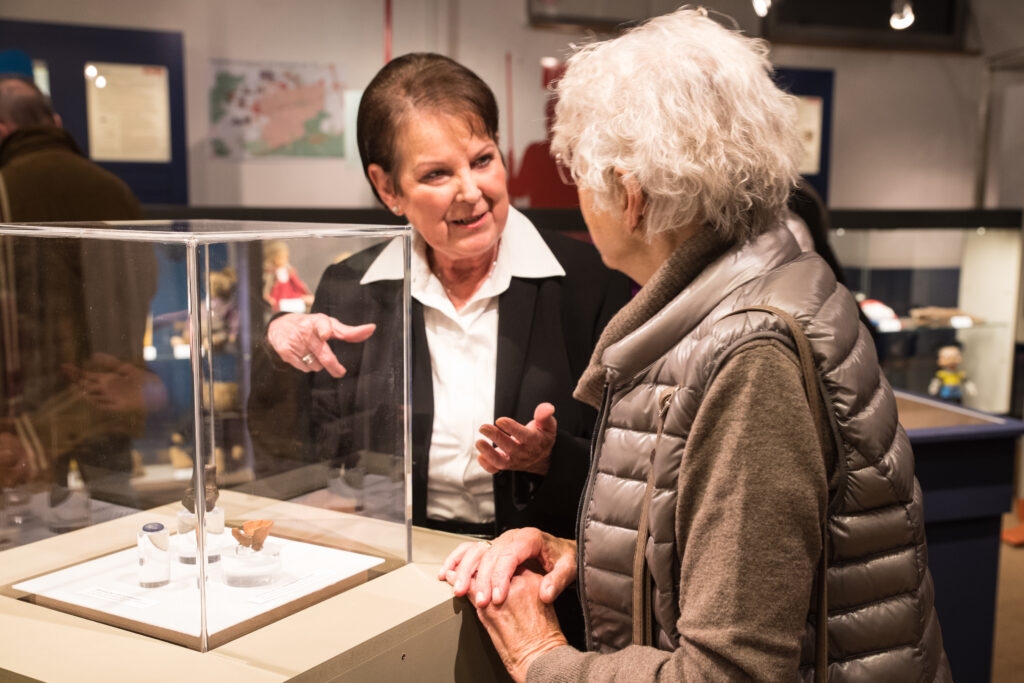 Sylvia Doll vom Vorstand des Fördervereins Stadtmuseum Germering im Gespräch mit einer Besucherin während der Spielzeugausstellung im Museum ZEIT+RAUM. Im Vordergrund eine Vitrine mit Ausstellungsstücken.
