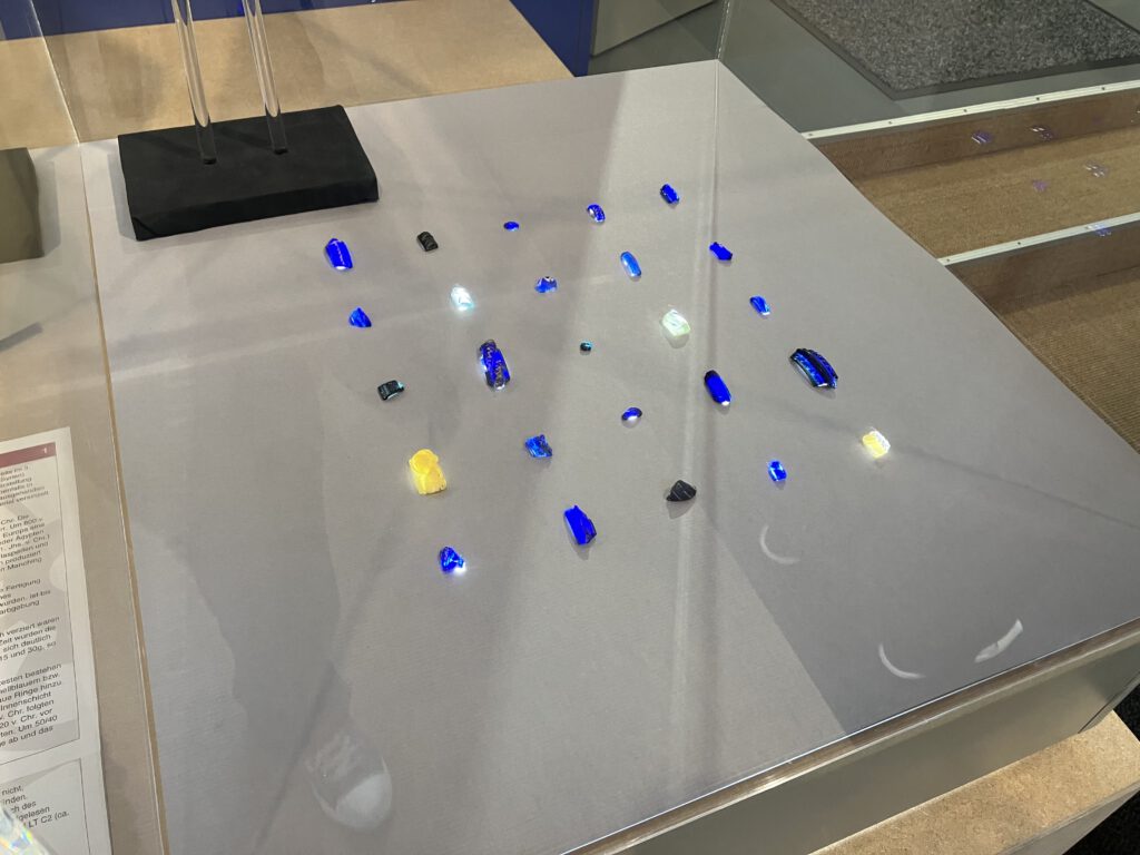 Leuchtend inszenierte Vitrine im Museum ZEIT+RAUM Germering mit farbigen Scherbenfunden, die von unten beleuchtet sind. Die Fundstücke in Blau, Gelb und Klarglas stammen vermutlich aus den Lesefunden.