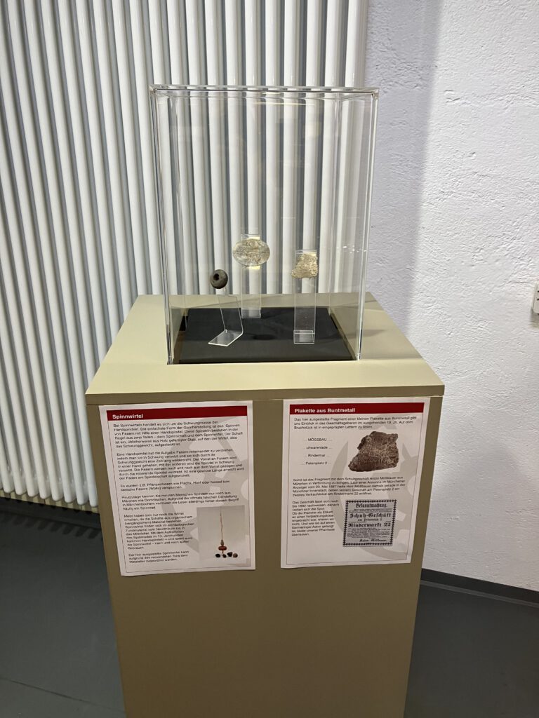 Ausstellungsvitrine im Museum ZEIT+RAUM Germering mit einem Spinnwirtel und zwei weiteren archäologischen Kleinfunden. Darunter zwei Infotafeln mit Erklärungen zu Spinnwirteln und einer Plakette aus Buntmetall.