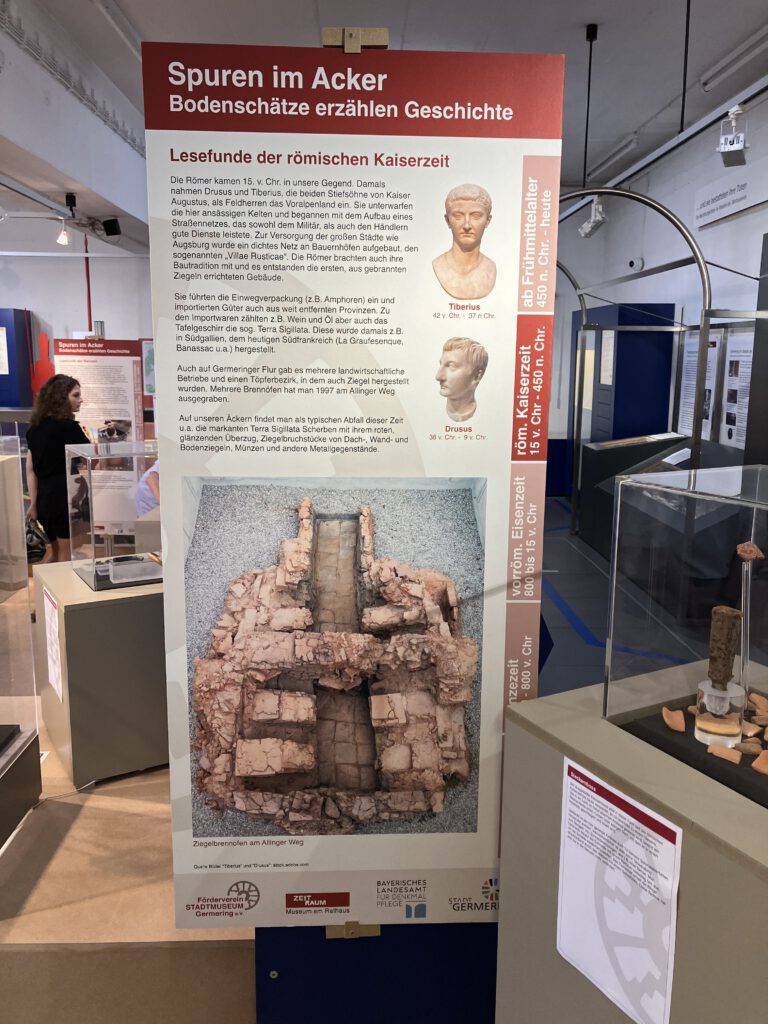 Infotafel zur römischen Kaiserzeit in der Ausstellung „Spuren im Acker“ im Museum ZEIT+RAUM Germering. Gezeigt werden Büsten von Tiberius und Drusus sowie ein Foto eines freigelegten Ziegelbrennofens. Der Text beschreibt Lesefunde wie Amphoren, Münzen und Terra Sigillata aus der Region.
