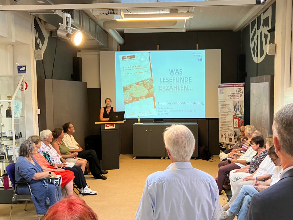 Eröffnung der Sonderausstellung „Spuren im Acker“ im Museum ZEIT+RAUM Germering am 19. Juli 2023. Dr. Sabine Mayer vom Bayerischen Landesamt für Denkmalpflege spricht am Rednerpult, vor ihr sitzt das Publikum. Im Hintergrund ist eine Leinwand mit dem Titel „Was Lesefunde erzählen…“ zu sehen.