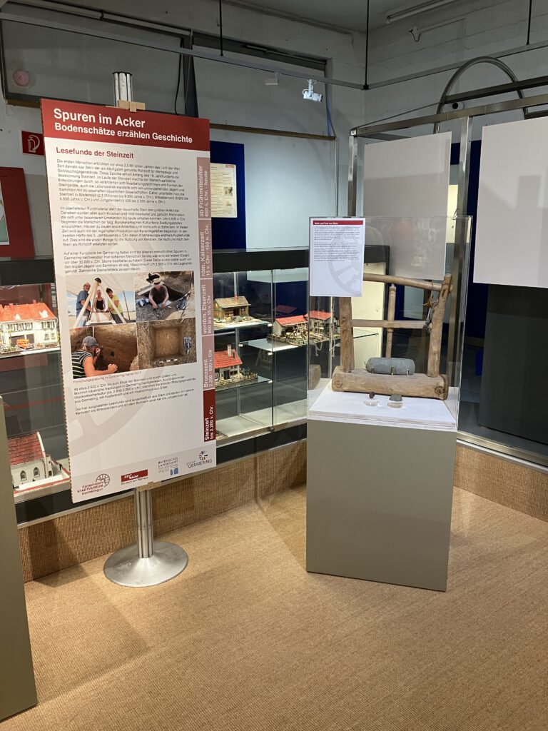 Ausstellungsvitrine im Museum ZEIT+RAUM Germering zur Sonderausstellung „Spuren im Acker“. Links eine Infotafel über Lesefunde der Steinzeit, rechts ein steinzeitliches Mahlwerk in einer Glasvitrine. Im Hintergrund Modelle historischer Gebäude.